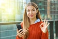 Close up of beautiful autumn woman holding smart phone doing Ok sign at the camera. Smiling cheerful satisfied young woman using