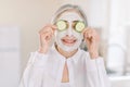 Close up of beautiful retired woman with facial mask on her face holding slices of fresh cucumber covering her eyes, on
