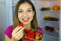 Close up of beautiful smiling girl near the fridge holding strawberries in her hands looks at camera. Healthy food concept. Fresh