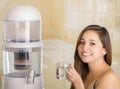 Close up of a beautiful smiling woman holding a glass of water, with a filter system of water purifier on a kitchen