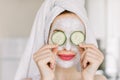 Close up of beautiful young woman with facial mask on her face holding slices of fresh cucumber covering her eyes, on Royalty Free Stock Photo