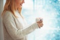 Close up of beautiful young woman in sweater holding cup of hot cocoa or coffee with marshmallow while standing near window indoor Royalty Free Stock Photo