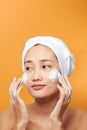 Close up beauty portrait of a laughing beautiful half naked Asian woman applying face cream and looking away isolated over orange Royalty Free Stock Photo