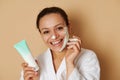 Young woman with beautiful toothy smile applying a facial cream or scrub on her face standing isolated over beige background with Royalty Free Stock Photo