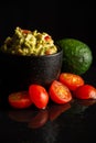 Close up of black bowl with guacamole on wet table with reflection with small tomatoes and avocado, selective focus,