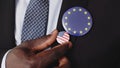 Close Up of Black Male Hand Compares American With European Union Flag Badge on Elegant Suit. Economy and Politics