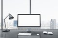 Close up of blank white computer screen on gray office desktop with objects, lamp and supplies on panoramic window and city view