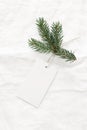 Close-up of blank white paper gift tag with rope and green fir branch on white linen tablecloth background. Price tag