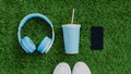 Blue wireless headphones, smartphone, cup of juice with sports sneakers on a green grass background, top view