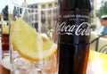Close up of a bottle of coca cola with text in spanish on a cafe table with a glass containing ice cubes and a slice of lemon