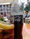 Close up of a bottle of coca cola with text in spanish on a cafe table with a glass containing ice cubes and a slice of lemon