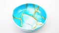 A close up of a bowl on a table. White and light cyan colors and cracks painted in gold, kintsugi