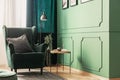 Close-up of a bright, green reading corner of a living room with a gray pillow on a velvet armchair next to a round, wooden coffee