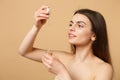 Close up brunette half naked woman 20s with perfect skin, nude make up apply oil from bottle on face isolated on beige