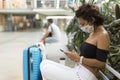 Close-up of a brunette woman using her smartphone while she sits next to her luggage. She`s wearing a face mask
