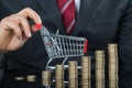 Close-up Of Businessman With Stack Of Coins And Shopping Cart