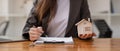 Close up businesswoman realtor taking notes and holding house model, sitting at desk with paper house model and keys