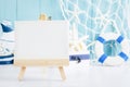 Close up of canvas and wooden easel with sail boat Toy model, ship wheel, rope and seashell on white and blue wooden background -