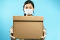 Close-up of a cardboard box in the hands of a young woman in a medical protective mask and gloves on a blue background Royalty Free Stock Photo