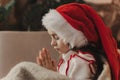 Close-up Caucasian child, lovely baby girl in Santa hat, makes cherished wish and cute presents, surrounded by Christmas
