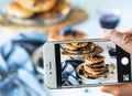 Close up of a cell phone taking a picture of a stack of blueberry pancakes against a blurred background of the scene.