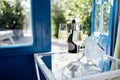 Close up of champagne glasses on transparent table. Bottle of wine and ice cooler. Blue door and garden with green trees on