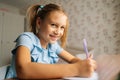 Close-up of cheerful elementary little child girl writing doing homework sitting at home table by window, looking at Royalty Free Stock Photo