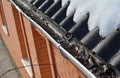 A close-up of a clogged, uncleaned roof gutter with last year fallen leaves and dirt while snow is melting on the roof of the