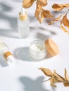 close up of collagen cream in glass jar, serum in white matte glass bottle and dry ruskus leaves on white background