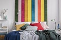 Close-up of a colorful bed with cushions and blankets standing against white wall with striped painting in bedroom interior. Real