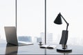 Close up of contemporary backlit workplace with laptop, table lamp, coffee cup and panoramic window with city view in the