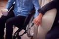 Close Up Of Couple At Home With Disabled Woman In Wheelchair Holding Hands With Man Sitting On Sofa