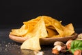 Close-up of crispy nachos on a a light wooden round plate. Corn chips next to nutsand basil leaves on a black background