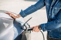 Close up cropped image of hands of man in jeans shirt and his electric car at charging station with the power cable Royalty Free Stock Photo