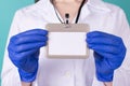 Close-up cropped photo of female doctor in white coat uniform gloves holding a blank mock up nametag in front isolated on blue