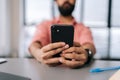 Close-up cropped selective focus shot of unrecognizable bearded man using smartphone sitting at office desk with book