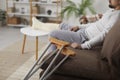 Close up of crutches leaned on the sofa, and a man with a broken leg sitting in the background Royalty Free Stock Photo