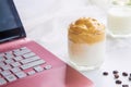 Close up a cup of Dalgona coffee with coffee beans next to a pink laptop on a white table with a cup of milk blurred background Royalty Free Stock Photo