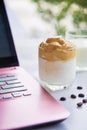 Close up a cup of Dalgona coffee with coffee beans next to a pink laptop on a white table with a cup of milk blurred background Royalty Free Stock Photo