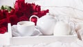 Close up of cup of tea, jar of milk, teapot on the wooden white tray with red roses nearby in the bed
