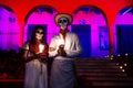 Close up of costumed man and woman with with mexican traditional white clothes and skull make-up in front of red pink lightened bu
