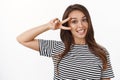 Close-up cute friendly-looking stylish young brunette in striped t-shirt showing peace, victory sign near eye smiling