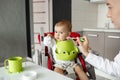 Close up of cute newborn son sitting in kitchen in baby chair and turning head aside refusing to eat baby food. Mother Royalty Free Stock Photo