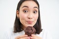 Close up of cute smiling asian woman, holding chocolate cupcake near mouth, having bite, enjoys eating pastry, white