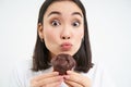 Close up of cute smiling asian woman, holding chocolate cupcake near mouth, having bite, enjoys eating pastry, white