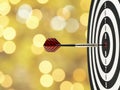 Close-up dart arrow hitting on target center on bullseye in wooden dartboard with blurred yellow gold lights bokeh background