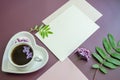 Close-up diagonal Mockup of white cards and a pink envelope on a dark purple background with leaves and lilac flowers with a coffe