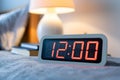 Digital clock shows 1200 on bedside table with blurred lamp and book Royalty Free Stock Photo