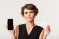 Close-up of disappointed, skeptical brunette woman smirk unamused, showing credit card and smartphone screen, standing