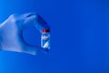 Close-up of a doctor`s hand with blue glove holding a vial of COVID-19 vaccine on blue background